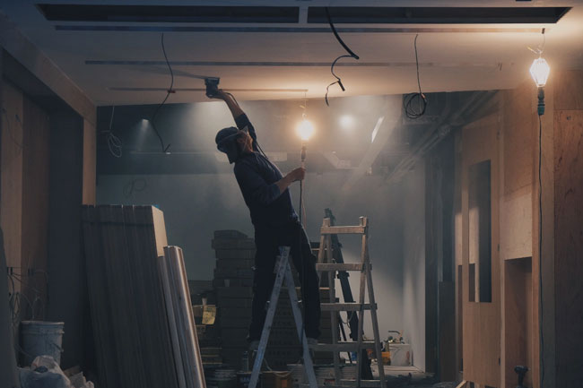 Un homme sur une échelle ponce un plafond. Il s'occupe de la rénovation d'un bâtiment commercial.